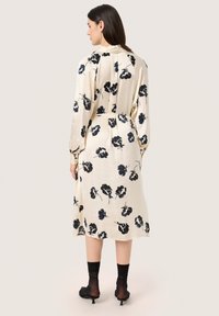 Rochie midi de culoare crem, cu un model floral negru, cu mâneci lungi, talie strânsă, fante laterale și un material moale și fin.