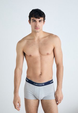Boxer-briefs en coton gris avec une ceinture navy arborant le logo Lacoste ; le modèle pose torse nu avec les bras le long du corps.