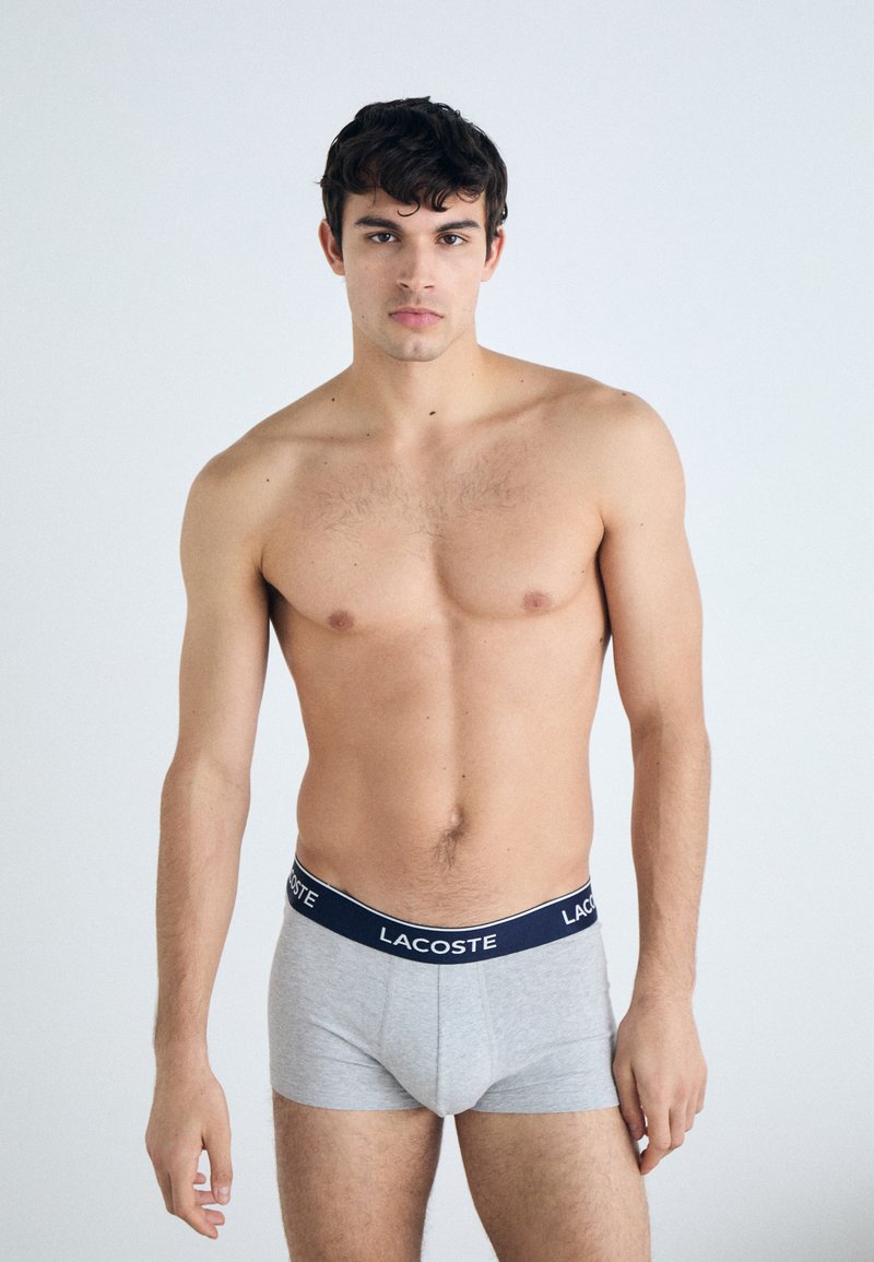 Graue Baumwoll-Boxershorts mit einem marineblauen Bund, der das Lacoste-Logo zeigt; das Modell posiert mit freiem Oberkörper und hängen gelassenen Armen.