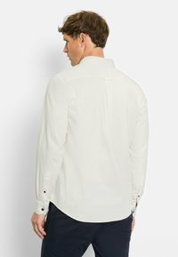 Langärmliges weißes Hemd aus leichtem Material. Verfügt über einen Standardkragen, Knopfmanschetten und eine Rückenfalte für zusätzliche Bewegungsfreiheit.