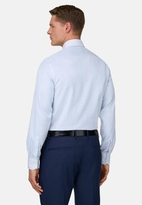 Mann, der rückwärts blickt, trägt ein hellblaues, langärmeliges Hemd, das in marineblaue Anzughose mit schwarzem Gürtel gesteckt ist.