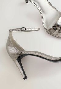 Metalické strieborné vysoké podpätky s hadím vzorom, špicatou špičkou a čiernym stiletto podpätkom; obsahujú nastaviteľný remienok na členku.