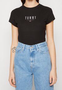 Zwarte katoenen T-shirt met korte mouwen, voorzien van een witte "TOMMY JEANS" logo, gecombineerd met hoge lichtblauwe spijkerbroeken.
