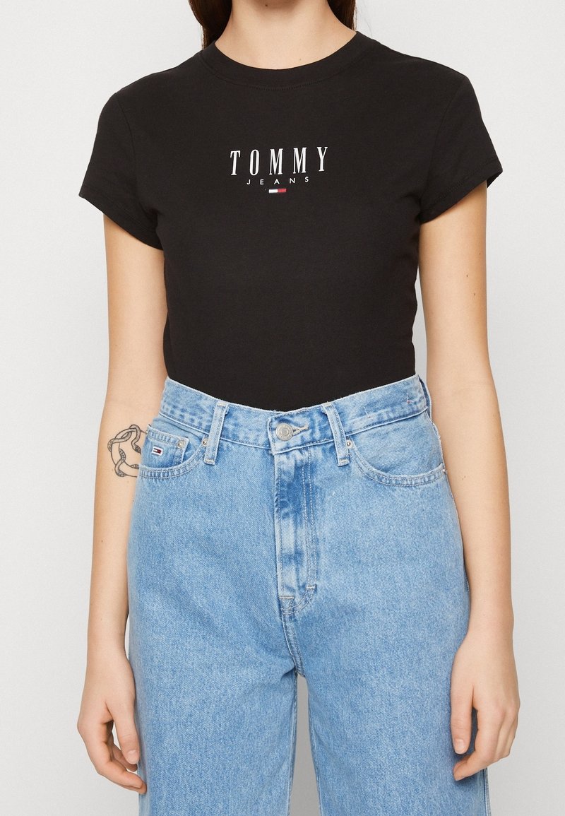 Zwarte katoenen T-shirt met korte mouwen, voorzien van een witte "TOMMY JEANS" logo, gecombineerd met hoge lichtblauwe spijkerbroeken.