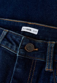 Tamnoplave traperice s bakrenim gumbom i kontrastnim narančastim šavovima. Oznaka glasi "TWO SOON," veličina 28/32.