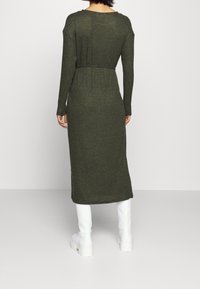 Lång, olivgrön stickad klänning med inriktad midja, långa ärmar och ribbad textur, parat med vita knä-höga stövlar.