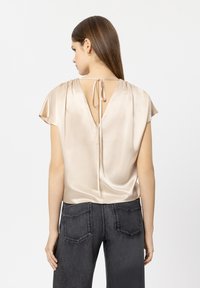 Satinbeige Bluse mit kurzen Ärmeln und tiefem V-Rücken, mit einer Schleifenbindung am Hals und einer glatten, glänzenden Textur.