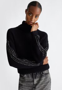 Maglione nero a maglia con colletto alto, caratterizzato da maniche texturizzate decorate con borchie argentate disposte in un motivo diagonale. Indossato con jeans grigi.