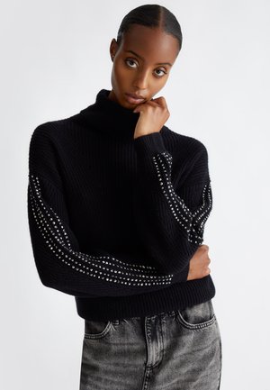 Maglione nero a maglia con colletto alto, caratterizzato da maniche texturizzate decorate con borchie argentate disposte in un motivo diagonale. Indossato con jeans grigi.