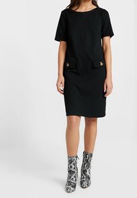 Černé šaty s krátkým rukávem a dvěma předními kapsami, se zlatými knoflíky, s rovnou siluetou. Nosené s bílými a černými kotníkovými botami se vzorem hada.