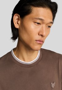 Maglietta a girocollo marrone con colletto bianco a costine e logo. Tessuto in cotone morbido, maniche corte e design aderente.