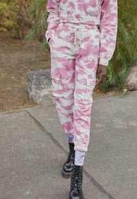 Calças de treino com padrão camuflado em branco e rosa, com bolsos laterais e punhos elásticos. Combinadas com botas pretas de atacador.