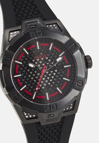 Reloj analógico negro con esfera texturizada, acentos rojos, marcadores de hora triangulares y correa de goma. Presenta manecillas de hora y minuto en negrita.