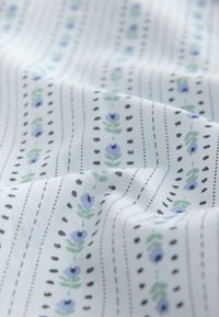 Puuvillakangas, jossa on valkoinen pohja ja toistuva kuvio sinisistä kukista, vihreistä lehdistä ja mustista pilkkuviivoista. Sujuva rakenne.