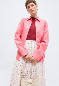 Γυναίκα που φοράει κοραλλί πουκάμισο με κουμπιά πάνω από μπορντό τοπ, λευκή φούστα με κόκκινο λουλουδάτο σχέδιο, κρατώντας μπεζ τσάντα με λεπτομέρεια φιόγκο.