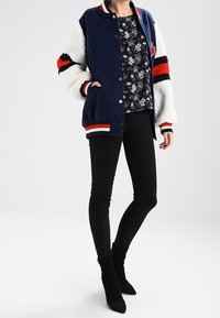 Veste universitaire bleu marine avec manches en polaire blanche, ornée de rayures rouges et noires. Portée sur un haut fleuri, associée à un jean skinny noir et des bottes.