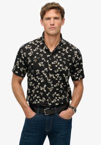 Camisa negra de manga corta con patrón floral blanco, de botones en la parte delantera, con dos bolsillos en el pecho, combinada con jeans oscuros y un cinturón negro.