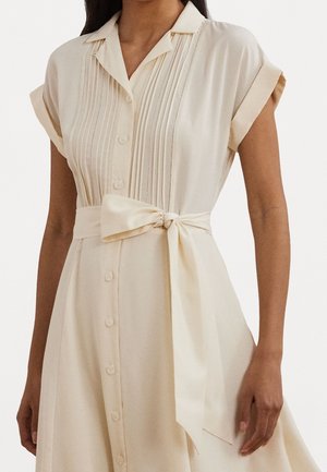 Robe beige boutonnée avec manches courtes retroussées, détails de plis verticaux sur le corsage et large ceinture nouée à la taille.
