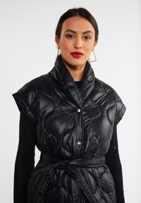 Gilet trapuntato nero con maniche corte, caratterizzato da un colletto alto, chiusure a bottone e una vita con cintura. Texture liscia con finitura lucida.