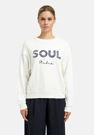 Frau mit hellbraunen Haaren in einem weißen Sweatshirt mit der Aufschrift "Soul Studio", kombiniert mit lockeren schwarzen Hosen, stehend vor einem einfarbigen Hintergrund.