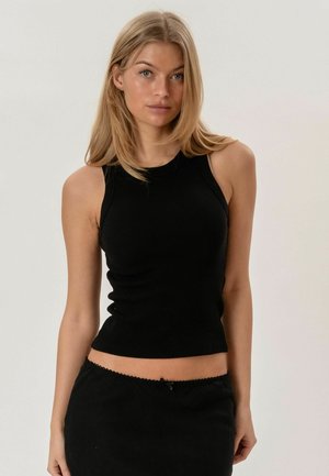 Jauna moteris su ilgais blondinais plaukais, vilkinti aptemptą juodą be rankovių palaidinę ir juodą sijoną ant paprasto šviesaus fono.