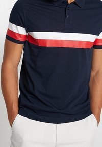 Polo de color navy hecho de algodón, con una franja horizontal roja y blanca en el pecho, cuello abotonado y mangas cortas.