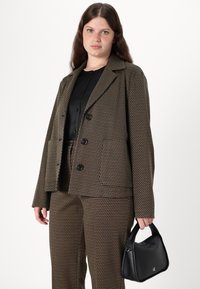 Blazer marron à motifs avec de grands boutons noirs, associé à un pantalon assorti. Un petit sac à main noir est tenu dans la main droite.