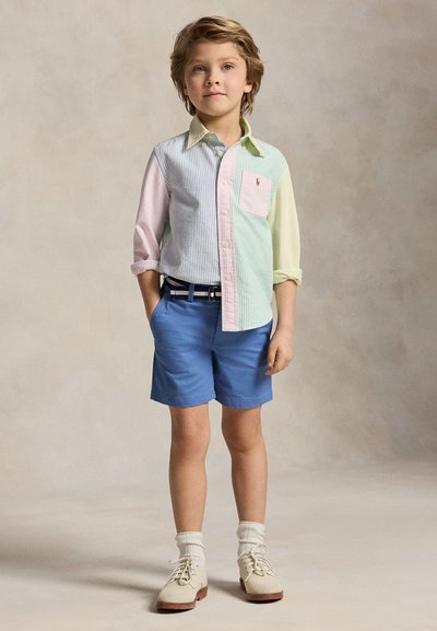 Νεαρός αγόρι στέκεται με τα χέρια στις τσέπες, φορώντας πουκάμισο με παστέλ ρίγες, μπλε σορτς, λευκές κάλτσες και μπεζ παπούτσια σε ουδέτερο φόντο.