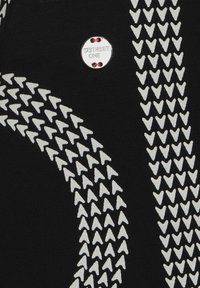 Svart tyg med vita pilmönster och ett litet runt metallmärke med ingraverad text "STREET ONE" fäst med röd tråd.