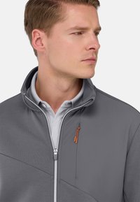 Veste grise zippée avec un col haut, présentant un tissu texturé et un tirette de fermeture éclair orange. Portée par-dessus un polo gris clair.
