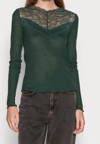Zelený top s dlouhým rukávem a krajkovým průhledem na výstřihu, se zubatými okraji a přiléhavým střihem. Kombinovaný s tmavými džíny.