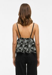 Top senza maniche nero con motivo floreale bianco, retro arricciato, spalline nere regolabili, indossato con pantaloni neri a vita alta.