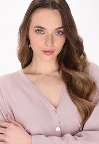 Cardigan rosa chiaro a V con maniche lunghe e tre bottoni frontali. Texture morbida con una leggera lucentezza. I capelli del modello sono mossi e sciolti.