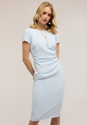 Blondynka w jasnoniebieskiej, dopasowanej, midi sukience z krótkim rękawem, nosząca złoty naszyjnik i duże perłowe kolczyki, na jednolitym tle.