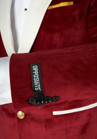 Roter Samtblazer mit kontrastierenden weißen Revers. Verfügt über schwarze Knöpfe, einen goldenen Akzent auf der Vorderseite der Jacke und ein farbenfrohes Einstecktuch.