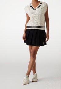 Chaleco de punto blanco con cuello en V y patrón de trenzas, combinado con una falda negra con pliegues. Zapatillas blancas con suela de goma completan el conjunto.