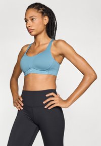 Sujetador deportivo de suave tela azul, con tirantes anchos y escote redondeado. Combinado con leggins negros de cintura alta para ropa deportiva.