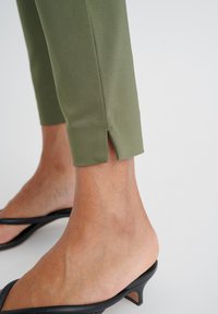Pantaloni tagliati verdi con una leggera apertura alla caviglia, abbinati a sandali aperti neri con cinturini. Tessuto liscio e design aderente.