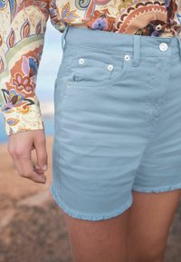 Vaaleansiniset farkkushortsit, joissa on repaleinen helma, etutaskut ja hopeasävyiset yksityiskohdat. Yhdistetty värikkään kukkakuosiset paitaan.