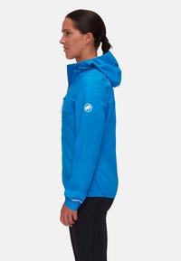 Giacca leggera blu con cappuccio, chiusura con zip e polsini elasticizzati. Presenta un logo sulla manica e una texture liscia e resistente all'acqua.
