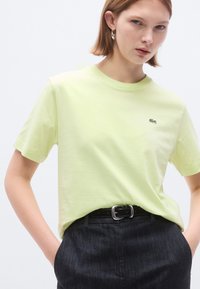 Ανοιχτοπράσινο βαμβακερό μπλουζάκι με στρογγυλή λαιμόκοψη, κοντά μανίκια και ένα μικρό κεντητό λογότυπο στο στήθος, συνδυασμένο με σκούρο παντελόνι και ζώνη.