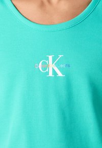 Turkizna rebrasta majica na naramenice z okroglim izrezom in belim logotipom "CK" na sredini prsi. Mehka tkanina z detajli teksture.