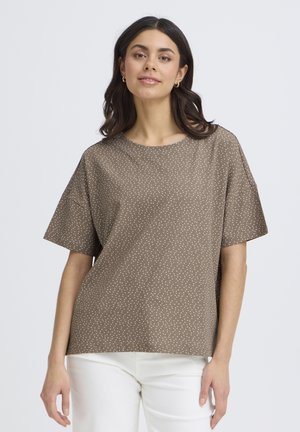 Femme aux cheveux foncés portant une chemise marron à manches courtes avec des pois blancs et un pantalon blanc, debout devant un fond uni.