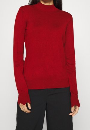 Femme portant un pull en maille rouge à manches longues avec des poignets étendus et un pantalon noir, posant devant un fond clair uni.