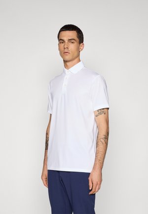 Man with tattoos wearing a white short-sleeve polo shirt and dark blue pants against a plain light gray background.