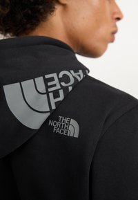 Felpa nera con cappuccio caratterizzata da un grande logo grigio sul retro e un logo più piccolo sulla parte anteriore a sinistra. Il materiale sembra morbido e con una texture.