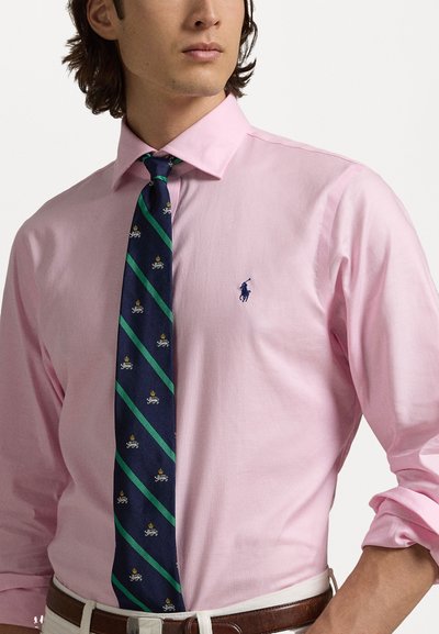 Camisa botão rosa clara com um pequeno logótipo azul no peito, combinada com uma gravata azul-marinho com riscas verdes e padrões bordados.