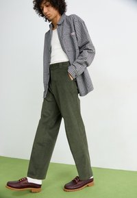Zwart-wit geblokte overhemd, witte tanktop, olijfgroene hoge broek en bruine schoenen met een dikke zool. Neutrale achtergrond.