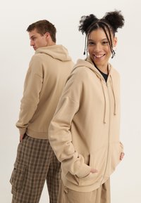 Two young adults wearing beige hoodies and casual pants, one smiling facing forward, the other turned away, standing against a plain background.