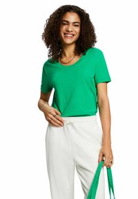 Camiseta verde de manga corta hecha de tela suave, con un cuello redondo. Combinada con pantalones blancos con cordón y sosteniendo tirantes verdes.